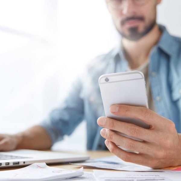 Modern smart phone in man`s hands. Young hipster guy with beard monitors news for website uses mobile phone, free internet and laptop computer. Skilled male journalist messages with chief editor